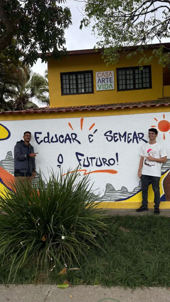O autor do blog e o artista Design do John posam em frente ao muro renovado da Casa Arte Vida. O muro branco possui um grafite colorido com a frase 'Educar é semear o futuro!' em letras azuis, acompanhado por desenhos de sol e montanhas.