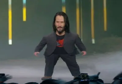 Meme de Keanu Reeves com o corpo digitalmente encolhido em proporções cômicas, parado no palco durante uma apresentação de videogame.