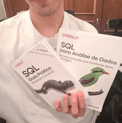 Close-up do autor segurando dois livros da O'Reilly sobre SQL. O livro à esquerda é "SQL Guia Prático" e o à direita é "SQL para Análise de Dados", focando em técnicas avançadas.